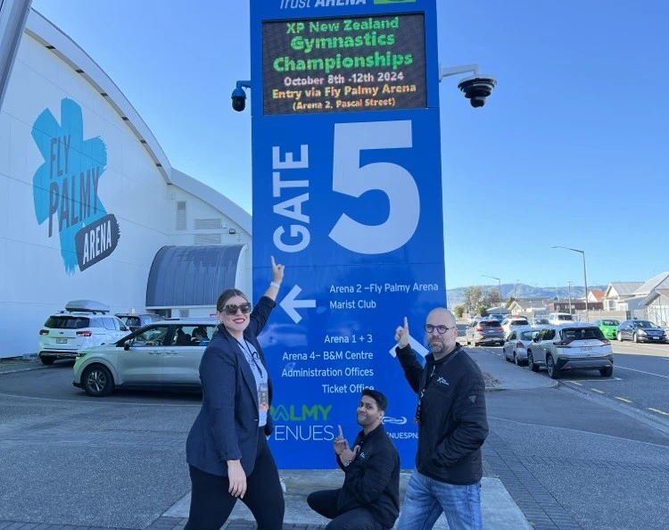 Group of people smiling and posing together at the XP Gymnastics Championships 2024, standing in front of a sign that reads XP Gymnastics Championships 2024 outside of Fly Palmy arena